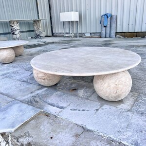 May include: A light-colored, round coffee table with a stone top and three spherical stone legs. Another table with a similar design is visible in the background. The tables are made of stone and are suitable for indoor or outdoor use.