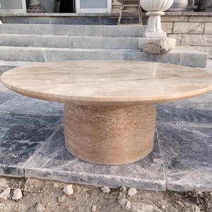 May include: A round, beige stone table with a cylindrical base. The table has a smooth, polished surface and is set on a stone patio. The table's diameter is approximately 48 inches.