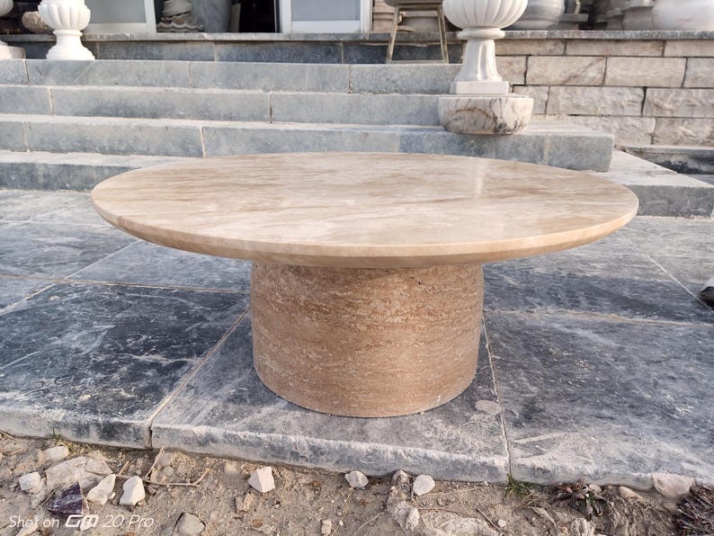 May include: A round, beige stone table with a cylindrical base. The table has a smooth, polished surface and is set on a stone patio. The table's diameter is approximately 48 inches.