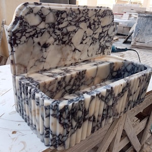 Puede incluir: Un lavabo rectangular de mármol con respaldo y frente acanalado. El lavabo es blanco con vetas grises oscuras. La parte trasera del lavabo tiene una parte superior curva. El lavabo está sobre una mesa de madera.