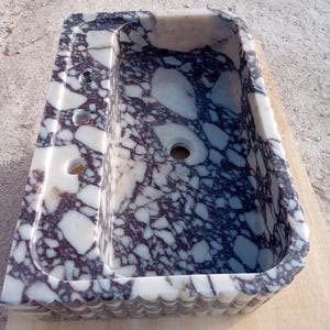 Puede incluir: Un lavabo rectangular de m&aacute;rmol con un dise&ntilde;o blanco y morado oscuro. El lavabo tiene un borde festoneado y tres agujeros en el lado izquierdo. El lavabo est&aacute; sobre una superficie de madera.