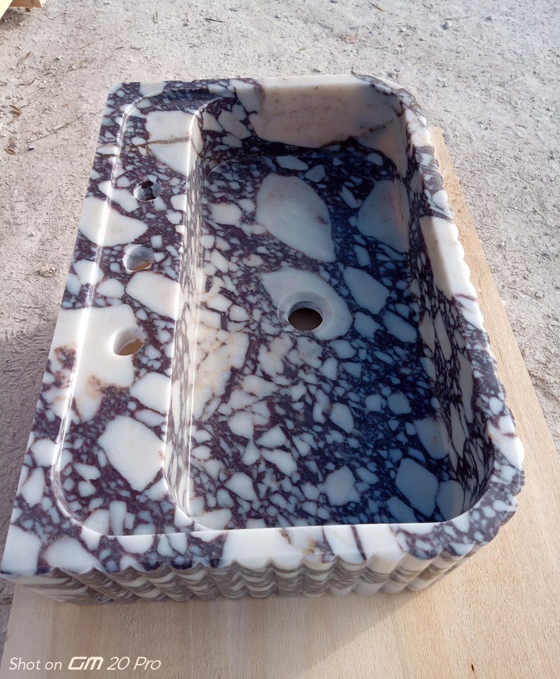 Puede incluir: Un lavabo rectangular de m&aacute;rmol con un dise&ntilde;o blanco y morado oscuro. El lavabo tiene un borde festoneado y tres agujeros en el lado izquierdo. El lavabo est&aacute; sobre una superficie de madera.