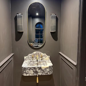 May include: A bathroom interior featuring a marble sink with gold fixtures, an oval mirror with a gold frame, and two glass sconces. The walls are painted a dark gray color, creating a modern and elegant aesthetic.