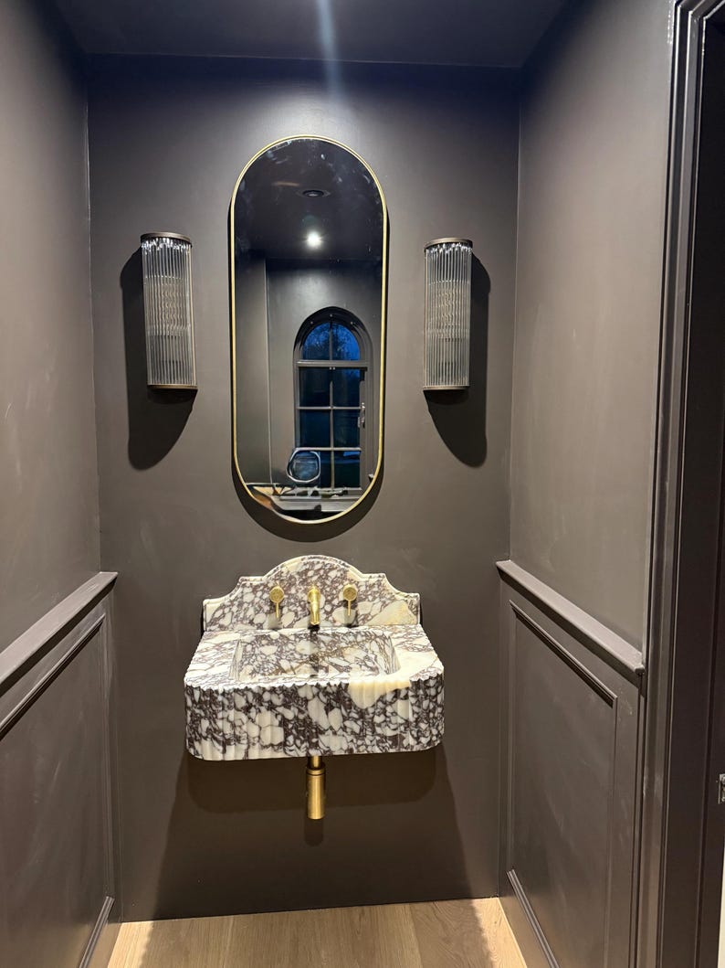 May include: A bathroom interior featuring a marble sink with gold fixtures, an oval mirror with a gold frame, and two glass sconces. The walls are painted a dark gray color, creating a modern and elegant aesthetic.