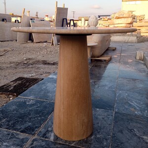 May include: A light brown, stone-like pedestal table with a round top. The table has a wide, conical base and a smooth, polished surface. The table's top is approximately 36 inches in diameter.