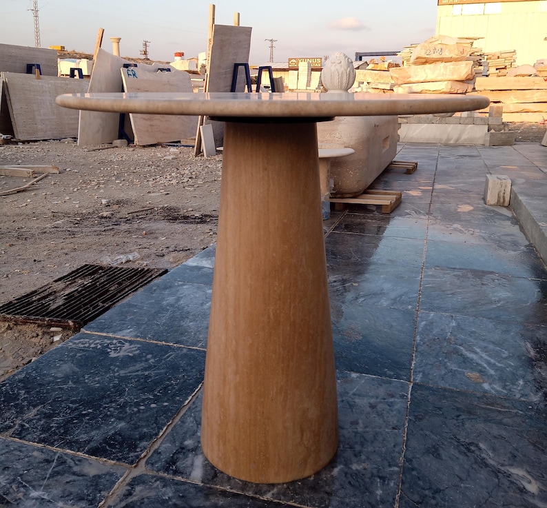 May include: A light brown, stone-like pedestal table with a round top. The table has a wide, conical base and a smooth, polished surface. The table's top is approximately 36 inches in diameter.