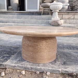 May include: A round, beige stone table with a cylindrical base. The tabletop is smooth and polished, while the base has a textured, layered appearance. The table is outdoors, set on a stone surface.