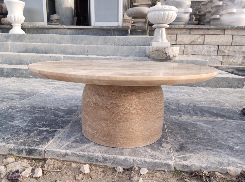May include: A round, beige stone table with a cylindrical base. The tabletop is smooth and polished, while the base has a textured, layered appearance. The table is outdoors, set on a stone surface.