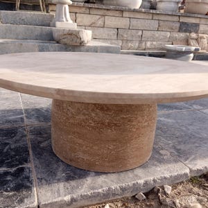 May include: A large, round outdoor table made of light brown stone. The tabletop is smooth and circular, supported by a cylindrical base. The table is set on a stone patio, with other stone structures in the background. The table is a neutral color.