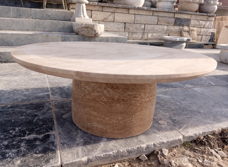 May include: A large, round outdoor table made of light brown stone. The tabletop is smooth and circular, supported by a cylindrical base. The table is set on a stone patio, with other stone structures in the background. The table is a neutral color.