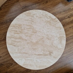 May include: A round, light-colored table top with a marbled pattern. The table top is made of a light beige material with darker beige and brown veining. The table top is set on a wooden floor.