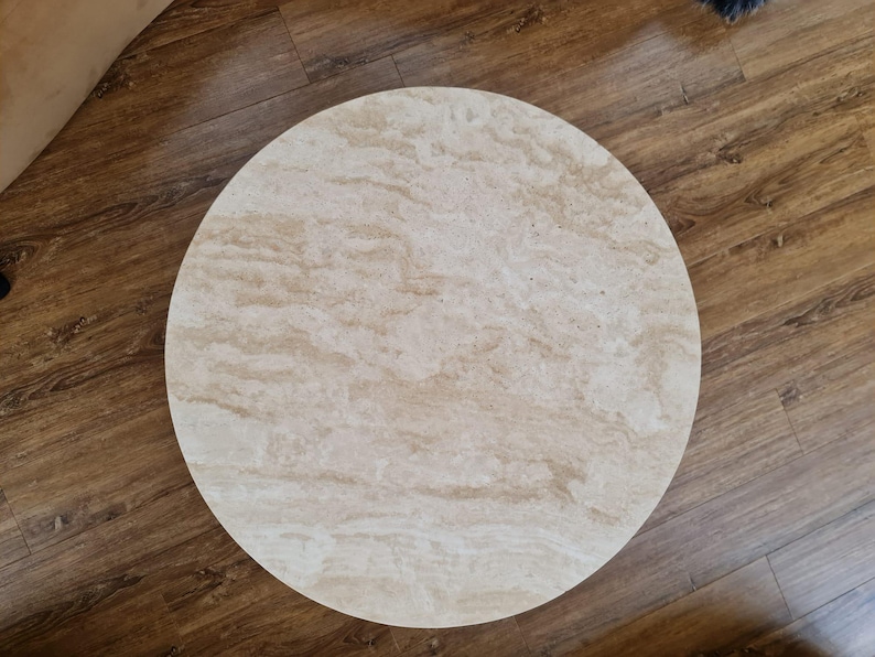 May include: A round, light-colored table top with a marbled pattern. The table top is made of a light beige material with darker beige and brown veining. The table top is set on a wooden floor.