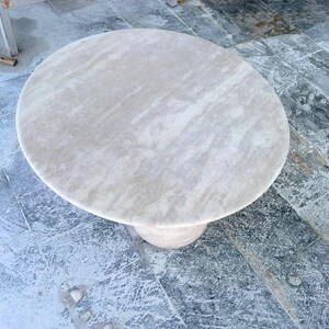 May include: A round, light-colored stone table with a single pedestal base. The tabletop has a smooth, marbled appearance with subtle variations in tone. The table is approximately 36 inches in diameter, suitable for a small dining area or as a decorative piece.