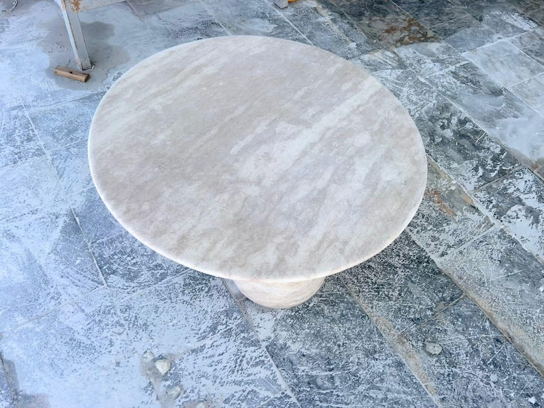 May include: A round, light-colored stone table with a single pedestal base. The tabletop has a smooth, marbled appearance with subtle variations in tone. The table is approximately 36 inches in diameter, suitable for a small dining area or as a decorative piece.