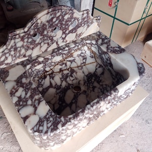 May include: A marble sink with a decorative backsplash. The sink basin is rectangular with a rounded front edge. The marble has a white and burgundy pattern. The backsplash has two holes for faucets.