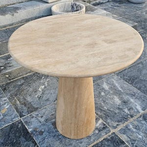 May include: A round, beige stone table with a single, cylindrical pedestal base. The tabletop has a smooth, polished surface. The table is made of a light-colored stone, likely travertine or marble, and is suitable for outdoor use.