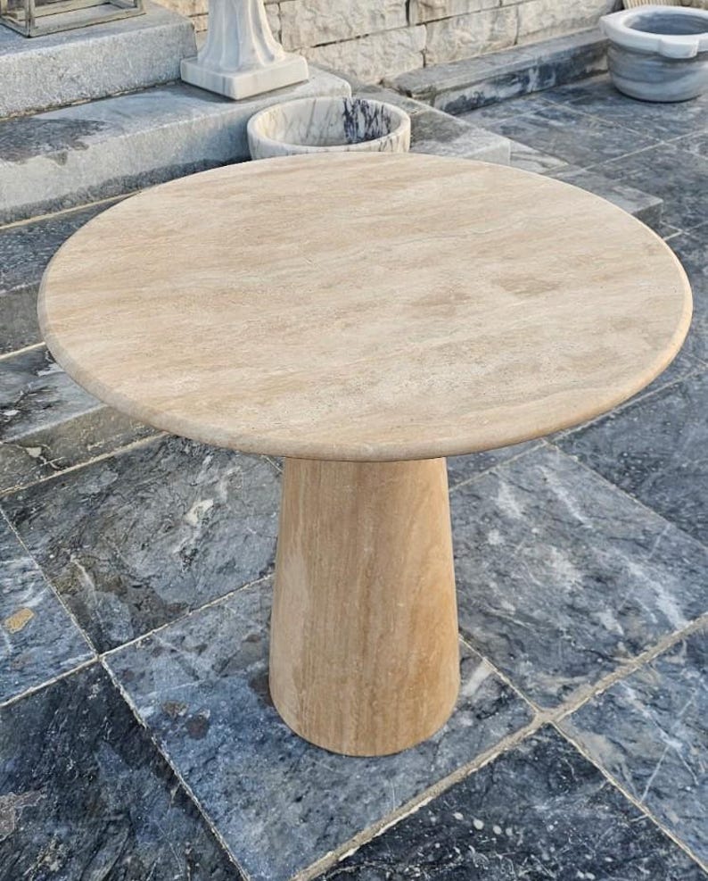 May include: A round, beige stone table with a single, cylindrical pedestal base. The tabletop has a smooth, polished surface. The table is made of a light-colored stone, likely travertine or marble, and is suitable for outdoor use.
