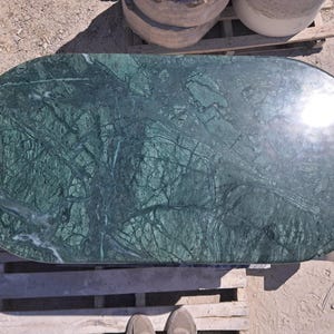 Puede incluir: Encimera ovalada de m&aacute;rmol verde pulido con intrincados patrones de vetas. La superficie de la piedra muestra una mezcla de tonos verdes oscuros y claros, creando un dise&ntilde;o natural y art&iacute;stico. La encimera est&aacute; sobre un pal&eacute; de madera.