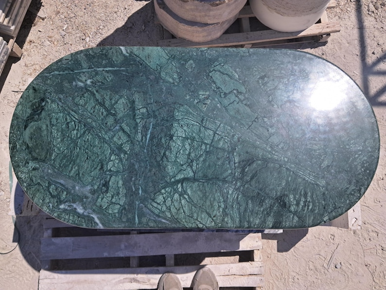 Puede incluir: Encimera ovalada de m&aacute;rmol verde pulido con intrincados patrones de vetas. La superficie de la piedra muestra una mezcla de tonos verdes oscuros y claros, creando un dise&ntilde;o natural y art&iacute;stico. La encimera est&aacute; sobre un pal&eacute; de madera.