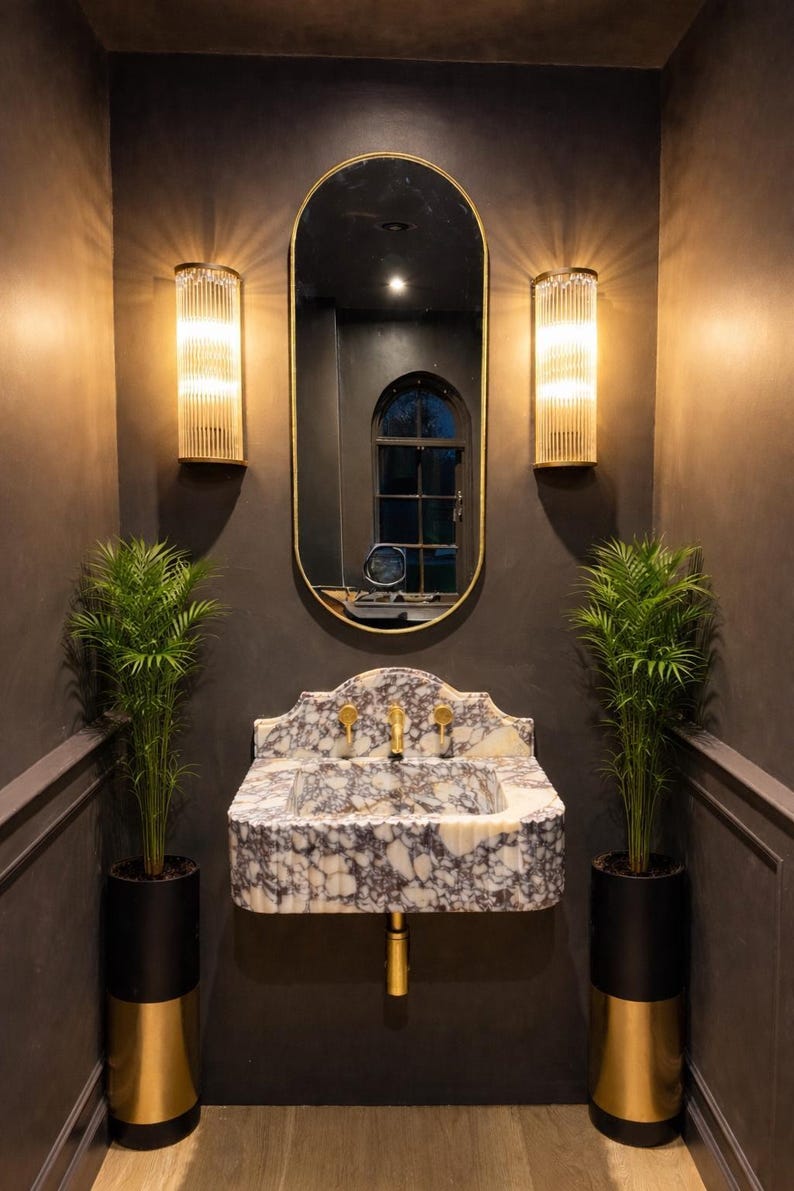 May include: A luxurious bathroom interior featuring a marble sink with gold fixtures, a large oval mirror, and two wall sconces. Two potted plants flank the sink, adding a touch of greenery to the space. The walls are painted a dark gray.