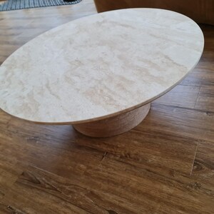 May include: Oval-shaped coffee table with a light beige marble top and a cylindrical base. The table is set on a hardwood floor with a warm brown tone. The design is modern and minimalist.