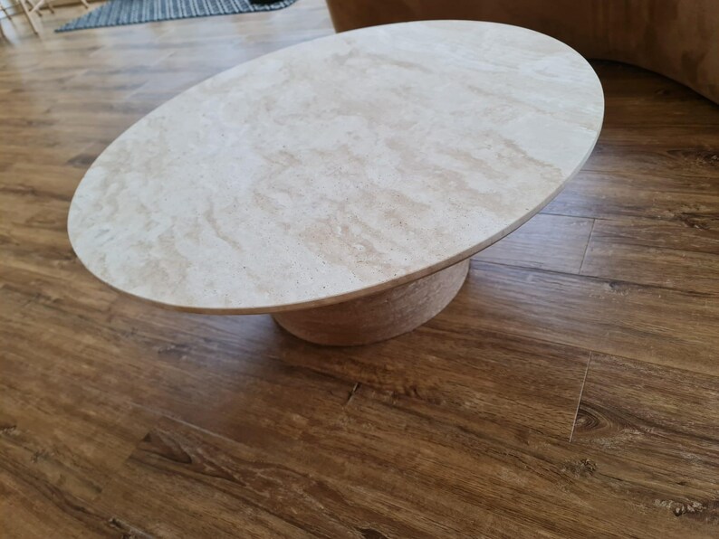 May include: Oval-shaped coffee table with a light beige marble top and a cylindrical base. The table is set on a hardwood floor with a warm brown tone. The design is modern and minimalist.
