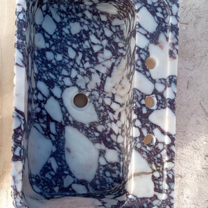 Puede incluir: Un lavabo rectangular de m&aacute;rmol con un patr&oacute;n &uacute;nico de vetas blancas y moradas oscuras. El lavabo tiene un orificio de drenaje central y tres orificios m&aacute;s peque&ntilde;os en el lado derecho. Los bordes tienen un dise&ntilde;o festoneado.