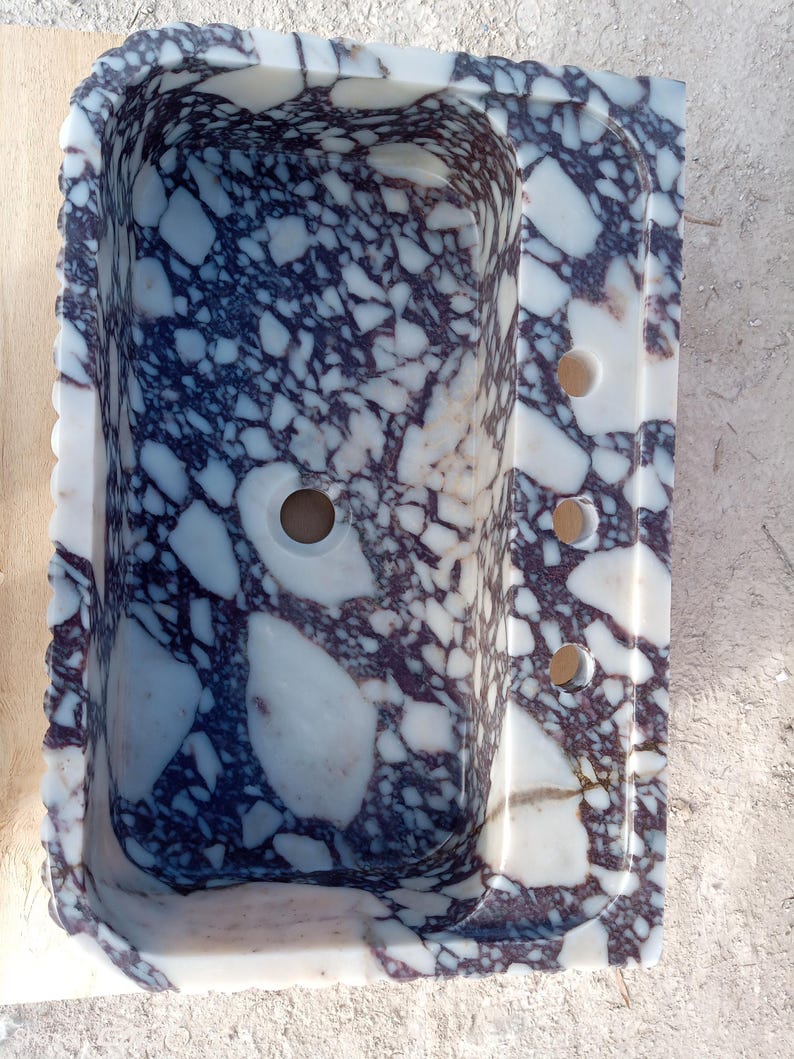 Puede incluir: Un lavabo rectangular de m&aacute;rmol con un patr&oacute;n &uacute;nico de vetas blancas y moradas oscuras. El lavabo tiene un orificio de drenaje central y tres orificios m&aacute;s peque&ntilde;os en el lado derecho. Los bordes tienen un dise&ntilde;o festoneado.
