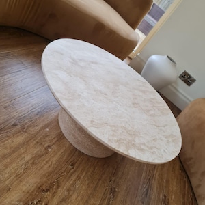 May include: A beige oval coffee table with a round base. The table is made of a light-colored stone material and sits on a wooden floor. The table's dimensions are approximately 48 inches long and 30 inches wide.