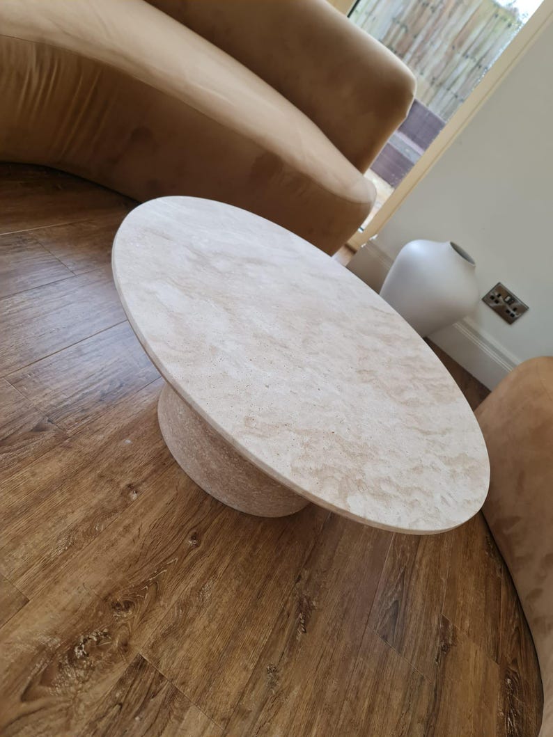 May include: A beige oval coffee table with a round base. The table is made of a light-colored stone material and sits on a wooden floor. The table's dimensions are approximately 48 inches long and 30 inches wide.