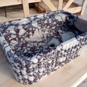 Puede incluir: Lavabo rectangular de m&aacute;rmol con un patr&oacute;n moteado blanco y morado oscuro. El lavabo tiene un dise&ntilde;o exterior acanalado y un orificio de drenaje. El lavabo est&aacute; hecho de piedra y est&aacute; sobre una superficie de madera.