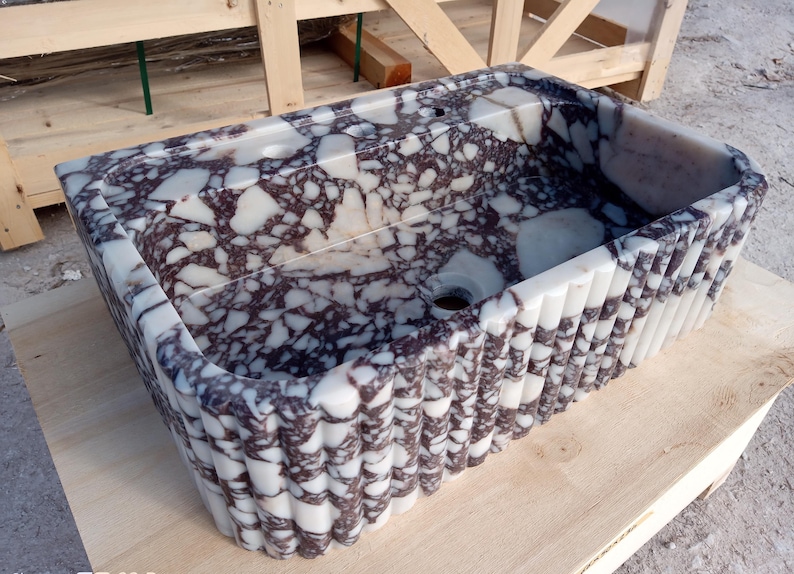 Puede incluir: Lavabo rectangular de m&aacute;rmol con un patr&oacute;n moteado blanco y morado oscuro. El lavabo tiene un dise&ntilde;o exterior acanalado y un orificio de drenaje. El lavabo est&aacute; hecho de piedra y est&aacute; sobre una superficie de madera.
