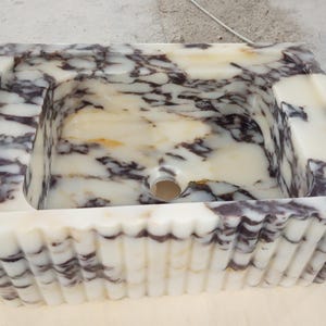 Puede incluir: Un lavabo rectangular de mármol con borde festoneado. El lavabo presenta una base blanca y crema con vetas moradas oscuras. El interior tiene un orificio de drenaje. Una adición única a cualquier baño.