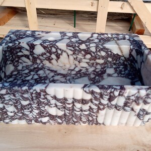 Puede incluir: Fregadero rectangular de m&aacute;rmol con interior estampado en blanco y morado oscuro. El exterior presenta un dise&ntilde;o acanalado. El fregadero es de piedra y se asienta sobre una superficie de madera.