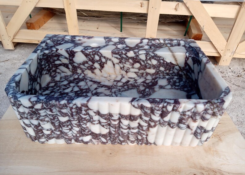 Puede incluir: Fregadero rectangular de m&aacute;rmol con interior estampado en blanco y morado oscuro. El exterior presenta un dise&ntilde;o acanalado. El fregadero es de piedra y se asienta sobre una superficie de madera.