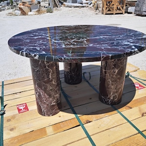 Pu&ograve; includere: Un tavolo rotondo in marmo con piano venato bordeaux scuro e bianco. Il tavolo ha tre gambe cilindriche. Il tavolo ha un diametro di circa 122 cm e un'altezza di 76 cm.