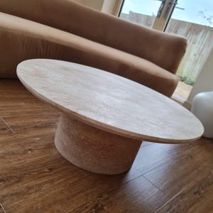 May include: Oval-shaped light wood coffee table with a cylindrical base. The table is approximately 40 inches long and 24 inches wide. The base has a textured, natural appearance. The table is set on a dark wood floor.