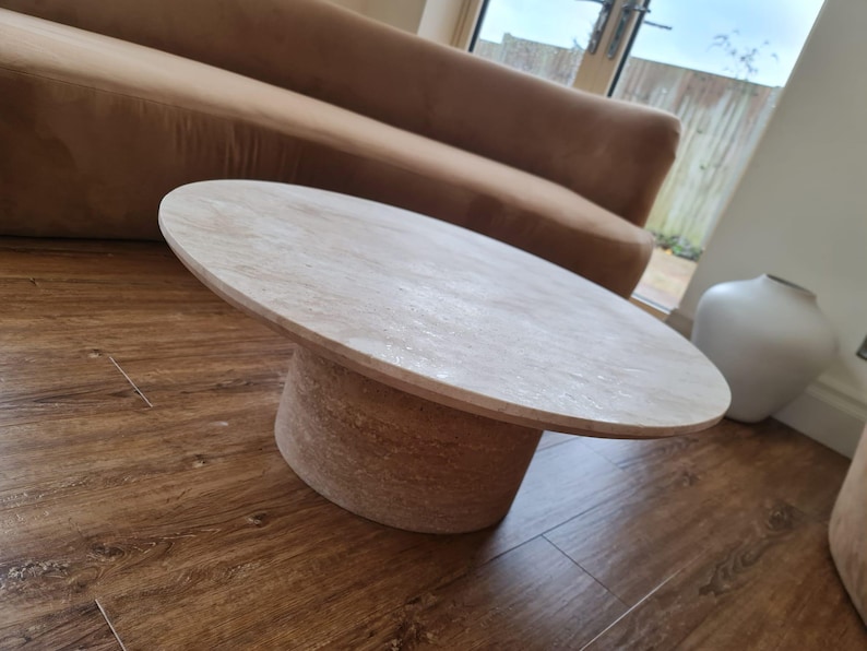 May include: Oval-shaped light wood coffee table with a cylindrical base. The table is approximately 40 inches long and 24 inches wide. The base has a textured, natural appearance. The table is set on a dark wood floor.