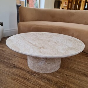 May include: A round, beige travertine coffee table with a cylindrical base. The table has a smooth, polished surface and is set on a dark wood floor. The background includes a brown sofa and a window.