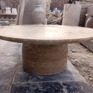 May include: A round, stone table with a light brown top and a cylindrical base. The table is made of a natural stone material and is suitable for outdoor use. The table's diameter is approximately 48 inches.