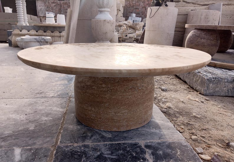 May include: A round, stone table with a light brown top and a cylindrical base. The table is made of a natural stone material and is suitable for outdoor use. The table's diameter is approximately 48 inches.