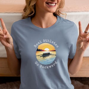 May include: A light blue t-shirt with a white graphic of a rowing crew in a boat on a lake at sunset. The graphic is surrounded by a circle with the text "Real Athletes Go Backward".