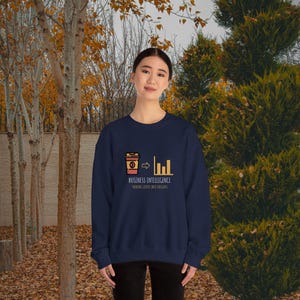 Op de afbeelding: Een marineblauwe sweatshirt met een afbeelding van een koffiekopje dat verandert in een staafdiagram, met de tekst "BUSINESS INTELLIGENCE" en "TURNING COFFEE INTO INSIGHTS". De sweatshirt wordt gedragen door een persoon.