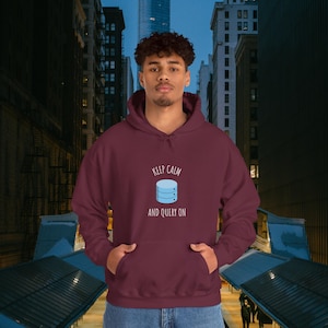 Op de afbeelding: Een bordeauxrode hoodie met een voorzak en een capuchon met trekkoord. De hoodie heeft een afbeelding van een database-pictogram met de tekst "KEEP CALM AND QUERY ON" in het wit. De achtergrond is een stadsgezicht.