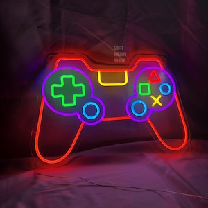 以下が含まれることがあります： ビデオゲームコントローラーの形をしたネオンサイン。 コントローラーは赤いアウトラインで、紫、緑、黄色、青のディテールがあります。 テキスト「GIFT NEON SHOP」が右上に表示されています。
