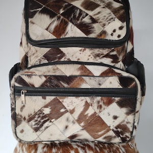 May include: A patchwork backpack with a geometric pattern of brown, white, and black cowhide squares. The backpack has a black leather trim, a zippered front pocket, and a top flap.