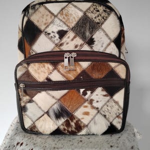 May include: A patchwork backpack and matching zippered pouch. The bags are made of diamond-shaped pieces of cowhide in shades of brown, black, and white, stitched together with white thread. The backpack has a large main compartment and adjustable straps.