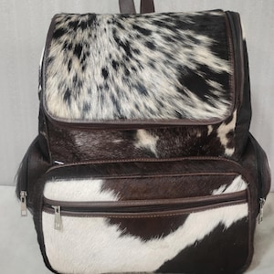 Könnte beinhalten: Ein Rucksack mit Kuhfellmuster in Schwarz, Weiß und Braun. Die Tasche verfügt über eine obere Klappe, eine vordere Reißverschlusstasche und seitliche Reißverschlussfächer, die alle mit dunkelbraunen Lederbesätzen und silberfarbenen Metallreißverschlüssen versehen sind.