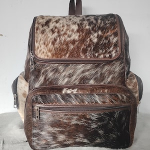 Puede incluir: Una mochila confeccionada en piel de vaca de color marrón, negro y blanco con un acabado texturizado y peludo. La bolsa cuenta con un compartimento principal grande con cremallera superior, un bolsillo frontal más pequeño y bolsillos laterales.