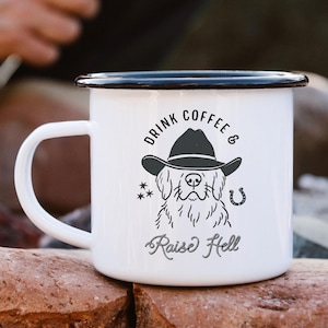 May include: White enamel camping mug with a black rim and handle. The mug features a graphic of a dog wearing a cowboy hat, with the text "DRINK COFFEE & Raise Hell". The mug is sitting on a rock.